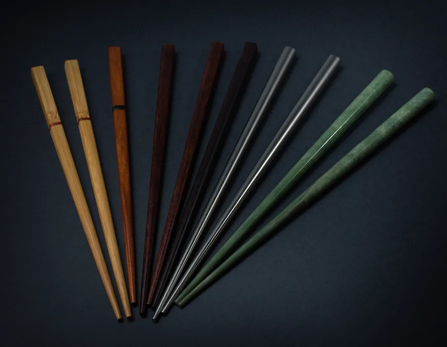 A fan of chopsticks made from many different materials including bamboo, dark wood, metal, and jade.