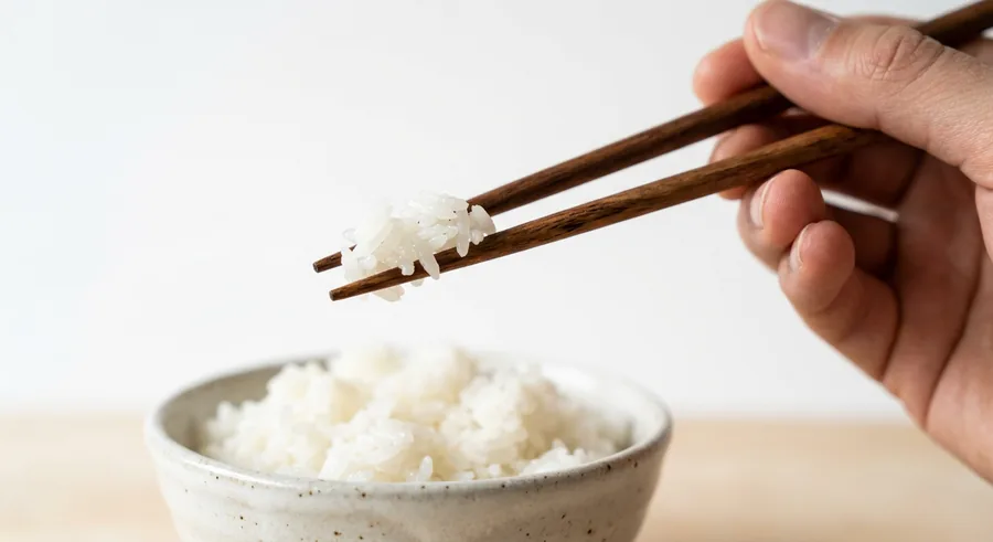 A confident hand using wooden chopsticks to lift a clump of sticky white rice from a ceramic bowl.